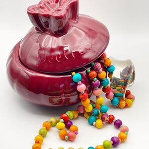 May include: A maroon ceramic jar with a flower-shaped lid, next to a colorful beaded necklace. The necklace features a variety of round beads in shades of pink, yellow, orange, green, and blue. The jar has the word "beet co" printed on it.