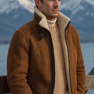 May include: A brown shearling jacket with a cream-colored shearling lining and collar. The jacket has a zip closure and is worn over a beige sweater. The background features a mountain range and a body of water.