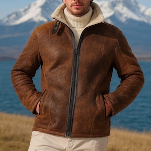 May include: Brown leather jacket with a cream-colored shearling collar and lining. The jacket has a zip-up front, two front pockets, and a black zipper. The person is wearing a cream-colored turtleneck sweater and khaki pants. The background features a mountain range and a body of water.