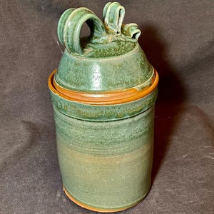 Puede incluir: Un tarro de cerámica hecho a mano con tapa. El tarro es cilíndrico con un esmalte verde y un borde marrón. La tapa tiene un asa decorativa. El tarro mide aproximadamente 15 cm de alto.