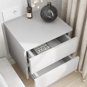 Puede incluir: Una mesita de noche gris moderna con dos cajones abiertos. La superficie superior contiene un jarrón gris oscuro, una pequeña botella marrón y flores blancas. El cajón superior contiene organizadores de joyas.