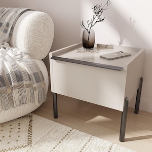 Puede incluir: Una mesita de noche moderna, de color crema, con patas de color gris oscuro y un asa a juego. Un jarrón con ramas decorativas, gafas y un smartphone descansan sobre la mesa. La mesita de noche está junto a un sillón color crema con una manta estampada.