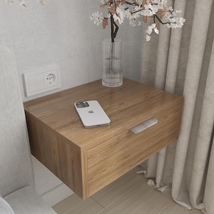 Puede incluir: Una mesita de noche flotante de madera con un cajón y un tirador plateado. Un smartphone descansa sobre la superficie, junto a un jarrón de cristal transparente con flores blancas. La mesita está montada en una pared gris claro.