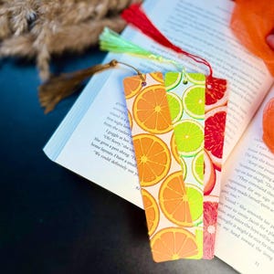 Puede incluir: Un conjunto de tres marcapáginas coloridos con diseños de frutas: rodajas de naranja, lima y pomelo. Cada marcapáginas tiene una borla. Los marcapáginas están colocados en un libro abierto.