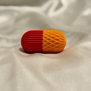 May include: A two-tone, pill-shaped object. One end is red with vertical ridges, and the other is yellow with a diamond-patterned texture. The object is resting on a white, silky surface.