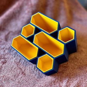 Pode incluir: Um organizador geométrico com design de favo de mel. O organizador é feito de plástico azul e amarelo. O interior de cada compartimento hexagonal é amarelo, enquanto o exterior é azul. Projetado para guardar pequenos objetos.