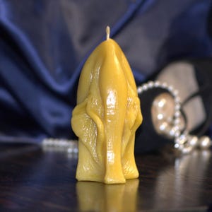 Puede incluir: Una vela de cera de abeja amarilla con un diseño esculpido único. La vela tiene una superficie texturizada y una mecha en la parte superior. La vela está sobre una superficie oscura, con un fondo azul y un collar de perlas.