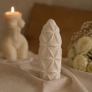 Może przedstawiać: Biała, cylindryczna świeca z powtarzającym się geometrycznym wzorem kwiatu życia wytłoczonym na powierzchni. Świeca ma spiczasty, stożkowy wierzchołek i jest wykonana z litego wosku, prezentując skomplikowane, symetryczne kształty.