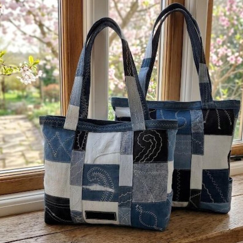 Pu&ograve; includere: Due borse tote in denim con design patchwork. Le borse presentano una variet&agrave; di tonalit&agrave; di denim blu, toppe bianche e nere, con cuciture a vista. I manici e i bordi superiori sono in denim pi&ugrave; scuro. Le borse sono su una superficie di legno.