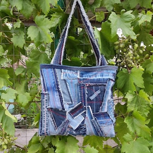 Op de afbeelding: Een draagtas gemaakt van verschillende tinten blauwe spijkerstof. De tas heeft een rechthoekig model met een patchworkontwerp, zichtbare gerafelde randen en rode decoratieve stiksels. De tas is voorzien van twee lange schouderbanden van denim.