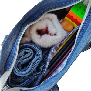 Pu&ograve; includere: Una borsa tote in denim blu con cerniera bianca, aperta per rivelare jeans arrotolati, un maglione bianco piegato con dettagli rossi, un cubo di Rubik e altri oggetti. La borsa &egrave; piena di effetti personali.