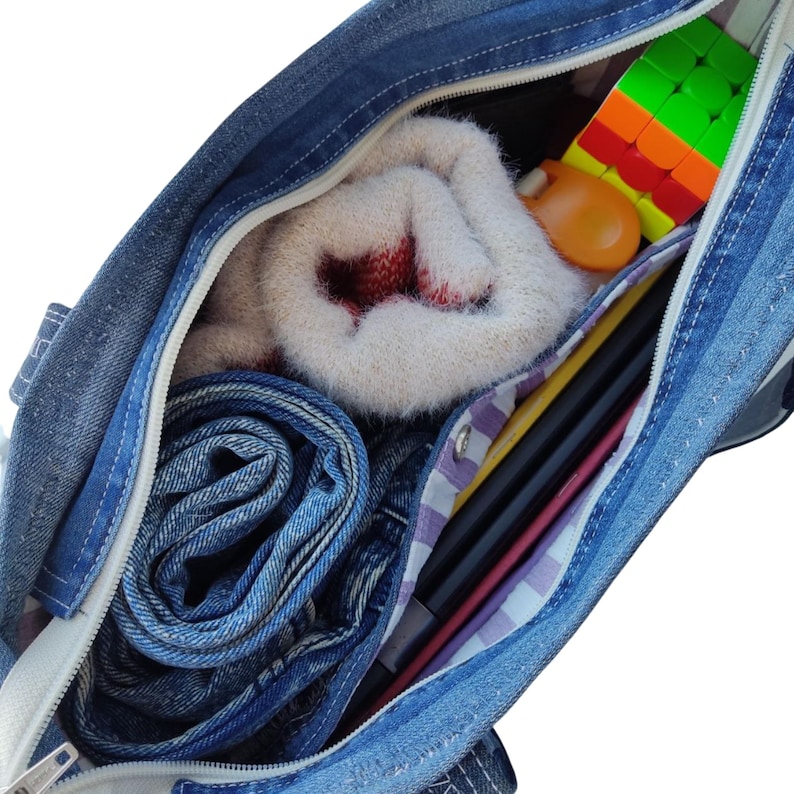 Pu&ograve; includere: Una borsa tote in denim blu con cerniera bianca, aperta per rivelare jeans arrotolati, un maglione bianco piegato con dettagli rossi, un cubo di Rubik e altri oggetti. La borsa &egrave; piena di effetti personali.