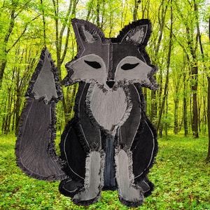 May include: A fox-shaped applique crafted from layered dark gray and light gray denim fabric pieces. The design features frayed edges along the seams, pointed ears, closed eyes, and a large bushy tail, creating a textured silhouette.