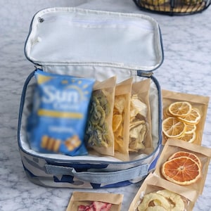 May include: A blue and white camouflage patterned lunch bag with an open lid, containing a blue snack bag and three clear pouches of dried fruit. Outside the bag are three additional clear pouches filled with dried strawberry, apple, and citrus slices.
