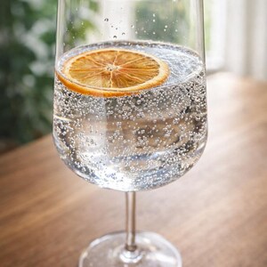 May include: A tall, clear wine glass filled with sparkling water and a slice of dried orange. The glass sits on a wooden surface. The drink is bubbly and refreshing, perfect for a summer day.