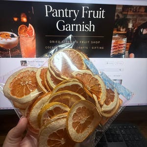 May include: A clear plastic bag filled with numerous dehydrated orange and grapefruit slices. The dried fruit rounds exhibit a translucent, textured appearance with visible pulp segments and rinds, showcasing shades of orange, yellow, and pink.