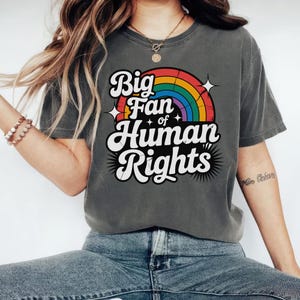 以下が含まれることがあります： 「Big Fan of Human Rights」の文字が白と黒で縁取られた、濃いグレーのTシャツ。テキストの上には虹のグラフィックがあります。カジュアルなスタイルのTシャツです。