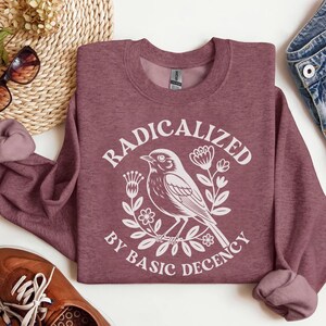 Może przedstawiać: Bordowa bluza z okrągłym dekoltem z białym napisem „RADICALIZED BY BASIC DECENCY”, otaczającym białą ilustrację ptaka i elementy kwiatowe. Bluza jest złożona na białej powierzchni.