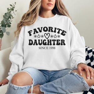 May include: White sweatshirt with the text "FAVORITE DAUGHTER" in black, with heart and star graphics, and the year "SINCE 1998". The sweatshirt is a crew neck style.