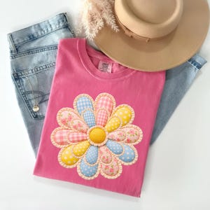May include: A pink t-shirt with a large daisy appliqué in pink, blue, and yellow patterns. The daisy has a yellow centre. A tan hat and blue jeans are also in the image.