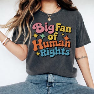 以下が含まれることがあります： レトロなフォントで「Big Fan of Human Rights」と書かれた、ダークグレーのTシャツ。 ヴィンテージ感のある、着古したような外観で、青いジーンズと合わせています。 デザインには星が含まれています。
