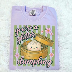Puede incluir: Una camiseta Comfort Colors lila claro con un gráfico que dice "you're a glitter dumpling". El gráfico presenta una empanadilla en una vaporera de bambú, flores rosas y rayas verdes.