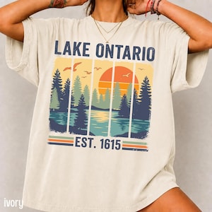May include: An ivory short-sleeve t-shirt featuring a graphic print with the text LAKE ONTARIO and EST. 1615. The design displays a sunset over a lake with pine trees and birds, divided into vertical panels with orange, blue, and green tones.