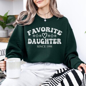 May include: A dark green sweatshirt with the words "FAVORITE DAUGHTER" in white, along with a heart and star graphic. The text "SINCE 1998" is below. The sweatshirt is a crew neck style.