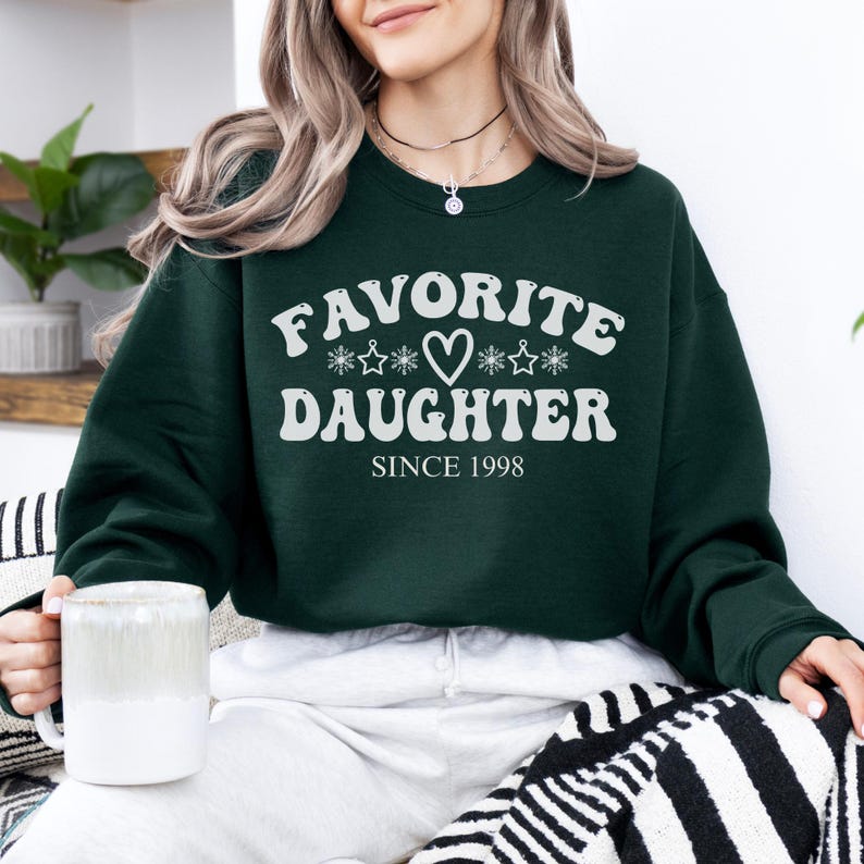 May include: A dark green sweatshirt with the words "FAVORITE DAUGHTER" in white, along with a heart and star graphic. The text "SINCE 1998" is below. The sweatshirt is a crew neck style.