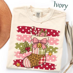 May include: An ivory t-shirt featuring a graphic of a Christmas stocking with a pink bow, a candy cane, and a gift box. The stocking has a leopard print section, a plaid pattern, and a gold bell. The background has horizontal polka dot stripes.