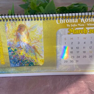 Puede incluir: Calendario de escritorio con espiral para marzo de 2026, con una ilustración de una mujer en un columpio. El calendario tiene un fondo amarillo con el texto "Chroma Kosmos Art" y "By Sofia Mata - Whimsical Art".