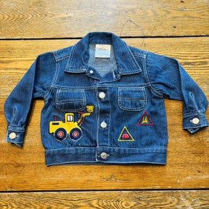 May include: A blue denim jacket with white stitching and button closures. The jacket features a yellow and red embroidered construction vehicle and road sign patches. The jacket has two chest pockets and buttoned cuffs. A small tag is visible at the collar.