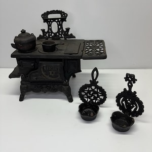 May include: A black cast iron toy stove with a kettle, pot, and two trivets. The stove has a decorative back and a burner grate. Two small pots are in front of the stove.