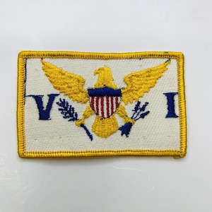 Op de afbeelding: Geborduurde patch met de vlag van de Maagdeneilanden. De rechthoekige patch heeft een witte achtergrond met een gouden rand. Het toont een gouden adelaar met een blauw en rood schild en de letters "V" en "I" in blauw.