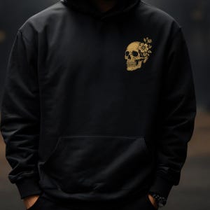 Puede incluir: Sudadera negra con un dise&ntilde;o de calavera dorada con detalles florales en el pecho izquierdo. La sudadera tiene un bolsillo delantero y una capucha con cord&oacute;n. El dise&ntilde;o combina elementos g&oacute;ticos y florales.
