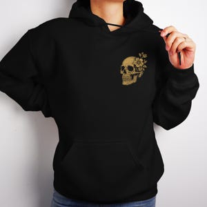 Puede incluir: Sudadera con capucha negra con bolsillo delantero y capucha con cord&oacute;n. El pecho superior izquierdo presenta un gr&aacute;fico de calavera dorada con un dise&ntilde;o floral. La calavera y la flor est&aacute;n detalladas con sombreado.