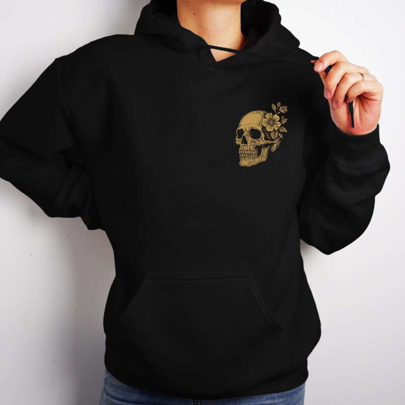 Puede incluir: Sudadera con capucha negra con bolsillo delantero y capucha con cord&oacute;n. El pecho superior izquierdo presenta un gr&aacute;fico de calavera dorada con un dise&ntilde;o floral. La calavera y la flor est&aacute;n detalladas con sombreado.