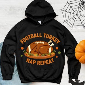 Puede incluir: Sudadera con capucha negra con las palabras "FOOTBALL TURKEY NAP REPEAT" en naranja. El diseño presenta un balón de fútbol americano y un pavo asado en una fuente. Un diseño divertido y festivo para el fútbol y Acción de Gracias.
