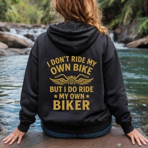 Puede incluir: Sudadera con capucha negra con texto dorado que dice "I DON'T RIDE MY OWN BIKE BUT I DO RIDE MY OWN BIKER" con un gr&aacute;fico de motocicleta. La sudadera con capucha la lleva una persona sentada cerca de una masa de agua.