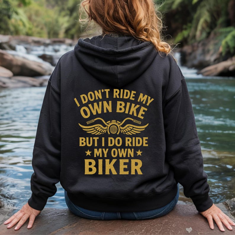 Puede incluir: Sudadera con capucha negra con texto dorado que dice "I DON'T RIDE MY OWN BIKE BUT I DO RIDE MY OWN BIKER" con un gr&aacute;fico de motocicleta. La sudadera con capucha la lleva una persona sentada cerca de una masa de agua.