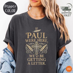 Puede incluir: Camiseta gris oscuro con el texto "IF PAUL WERE HERE, WE'D BE GETTING A LETTER." El diseño incluye una cruz, plumas y elementos decorativos dorados. La camiseta tiene mangas cortas.