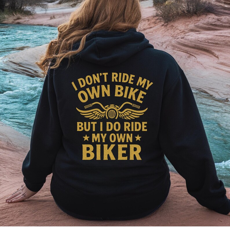 Puede incluir: Sudadera con capucha negra con texto dorado que dice "I DON'T RIDE MY OWN BIKE BUT I DO RIDE MY OWN BIKER" con un gr&aacute;fico de motocicleta. La sudadera la lleva una persona con pelo largo y casta&ntilde;o claro.