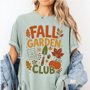 Puede incluir: Una camiseta de color verde salvia claro con un estampado gráfico que incluye el texto FALL GARDEN CLUB. El diseño presenta ilustraciones de un sobre de semillas, una pala de jardín, una bellota, hojas de otoño y una calabaza naranja.
