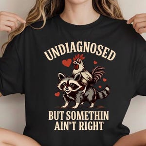Puede incluir: Camiseta negra con el texto "UNDIAGNOSED" sobre un dibujo de un mapache llevando un gallo, con el texto "BUT SOMETHIN AIN'T RIGHT" debajo. El diseño incluye corazones rojos.