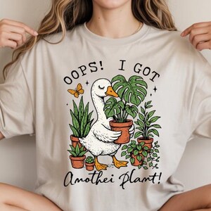 Puede incluir: Camiseta beige con una ilustración de un ganso sosteniendo una planta en maceta, rodeado de otras plantas en maceta y una mariposa. El texto dice "OOPS! I GOT Another Plant!" en una fuente juguetona.