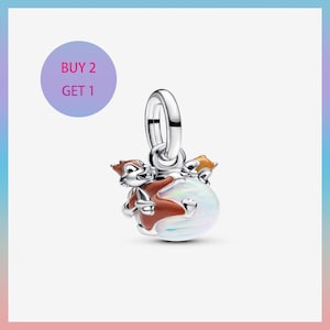 May include: A silver charm featuring a squirrel design. The squirrel is brown and white, clutching a nut, and is positioned on a white and iridescent orb. The charm has a silver loop for attachment. A purple circle with the text "BUY 2 GET 1" is in the background.