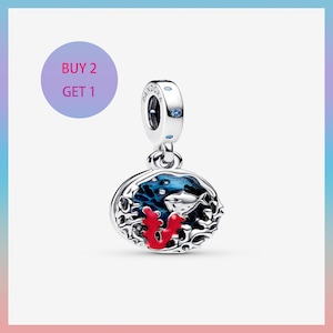 Puede incluir: Un charm de plata con una escena oceánica azul y roja. El charm presenta un pez, coral y un fondo de esmalte azul. El charm tiene un logotipo de Pandora y una gema azul. La imagen también incluye un círculo morado con el texto "COMPRA 2 OBTÉN 1".
