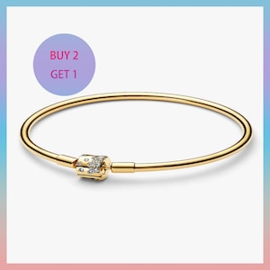 May include: A gold-colored bangle bracelet with a decorative clasp. The clasp is adorned with small, clear gemstones. The bracelet is displayed on a white background with a purple circle that says "BUY 2 GET 1".