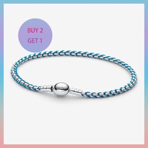 May include: A silver Pandora bracelet featuring a woven design with bright blue cord detailing. The bracelet includes a round, polished silver clasp with the Pandora logo engraved on the front. A circular purple graphic displays the text BUY 2 GET 1.
