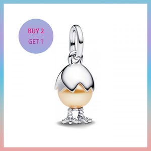 Puede incluir: Un charm de plata con forma de pollito saliendo de un huevo. El huevo es de color amarillo claro y las patas del pollito son visibles en la parte inferior. El charm tiene un lazo en la parte superior para sujetarlo a una pulsera o collar. El texto "BUY 2 GET 1" está en un círculo morado.